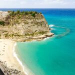 Spiaggia siciliana con sabbia dorata e acque cristalline, circondata da scogliere e vegetazione mediterranea.